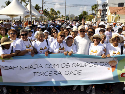 Centenas de idosos com camisetas brancas estão em uma caminhada. Os da frente seguram uma faixa onde se lê: caminhada com os grupos da terceira idade