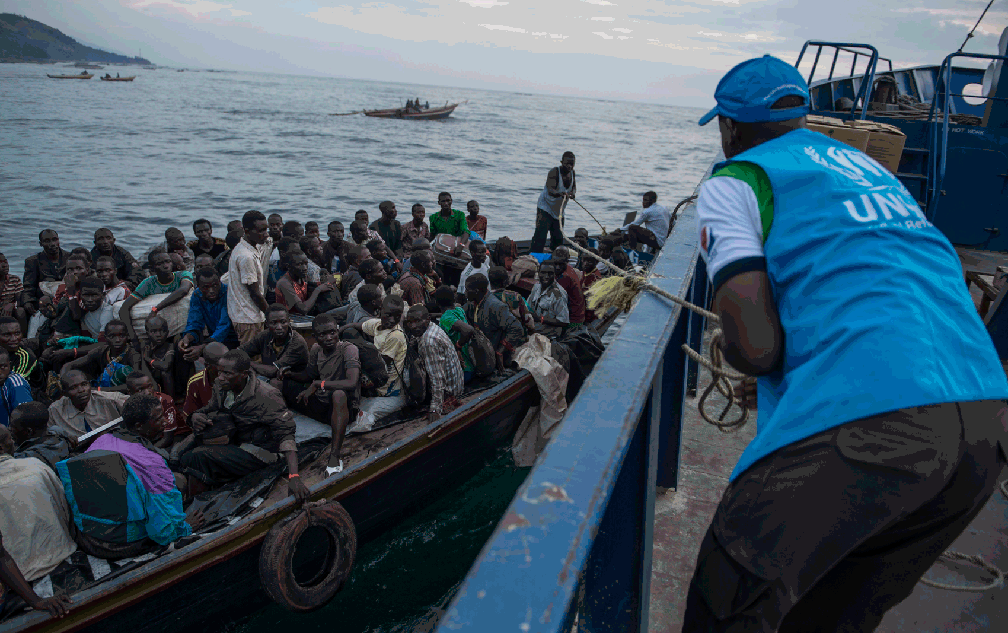 Homem de costas em primeiro plano, com colete da ACNUR, está em uma embarcação de onde vê um barco lotado de homens e mulheres